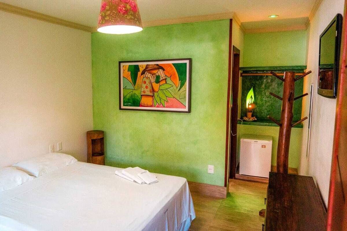 A hotel room with a bed, a painting on the wall, a wooden nightstand, and a small heater.