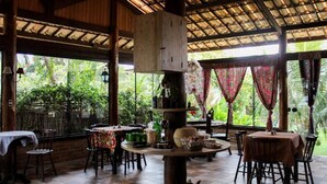 Un comedor interior con mesas y sillas de madera, una columna de madera en el centro, y vistas de exuberante follaje afuera.