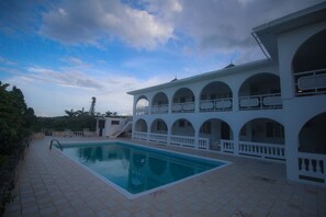 A swimming pool with a tiled deck, surrounded by a white building with arched balconies.