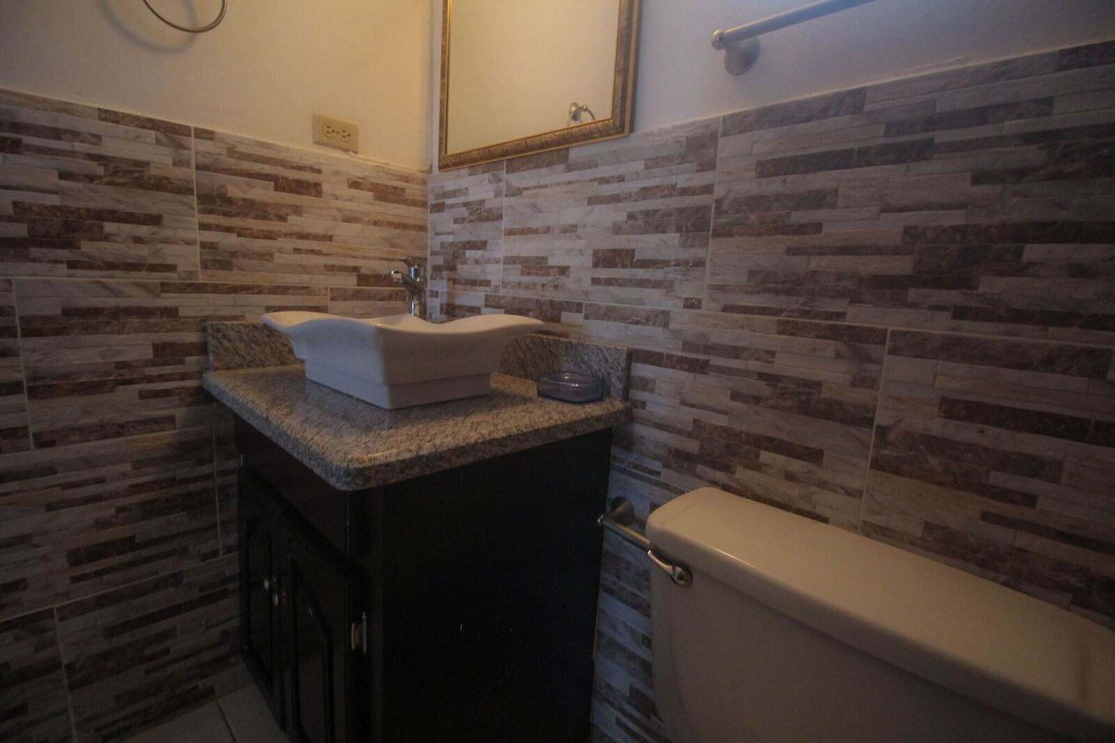 A bathroom with a granite countertop, a white sink, and a toilet. The walls are tiled with a patterned design.
