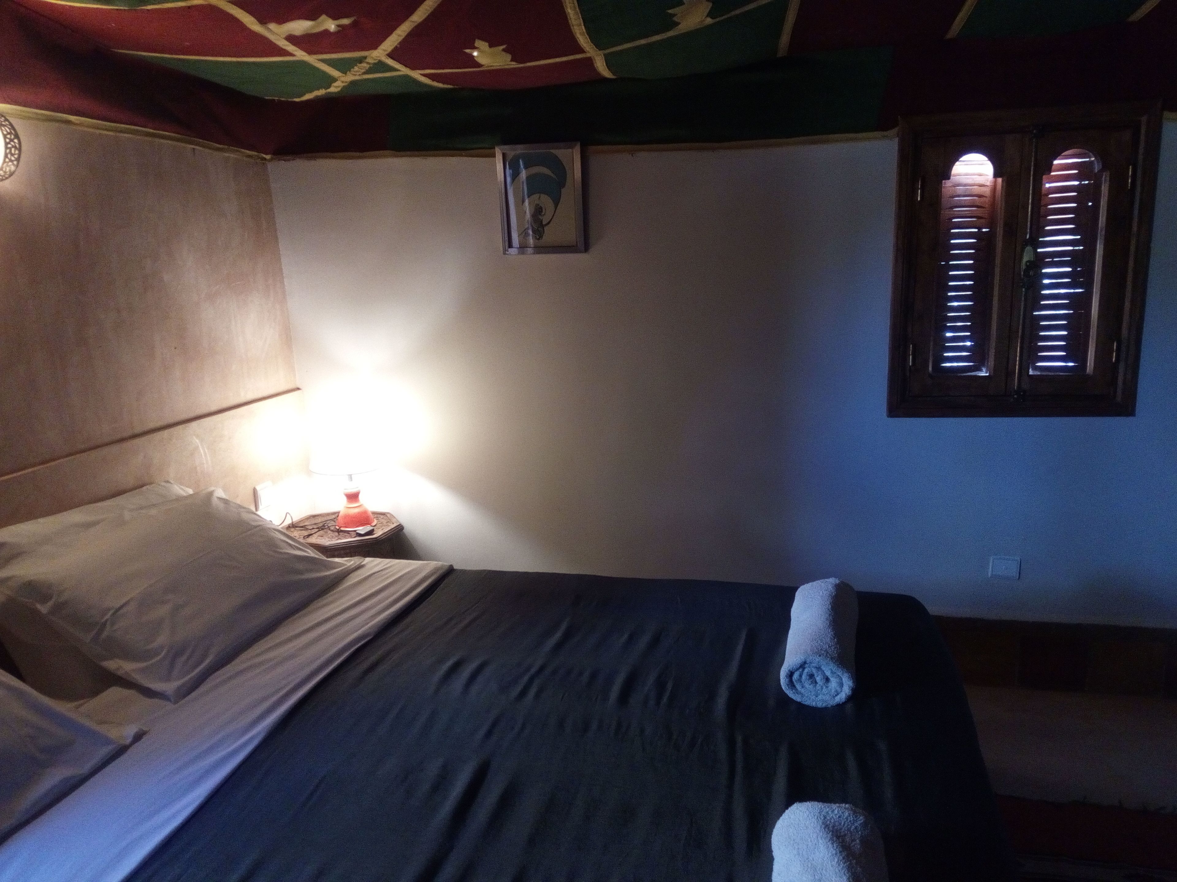 A room with a bed, a nightstand with a lamp, a window with blinds, and a wall-mounted picture.