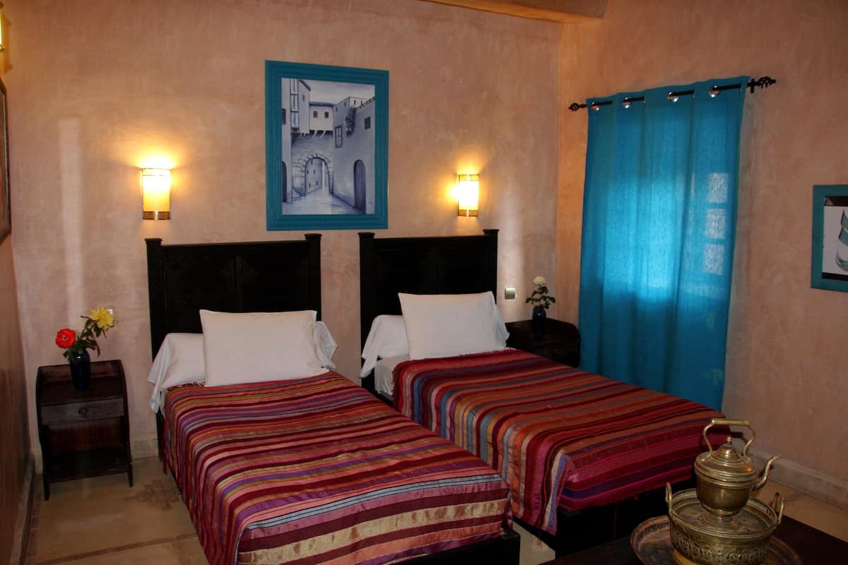 A hotel room with two beds, a nightstand with a vase of flowers, a painting on the wall, and a blue curtain.