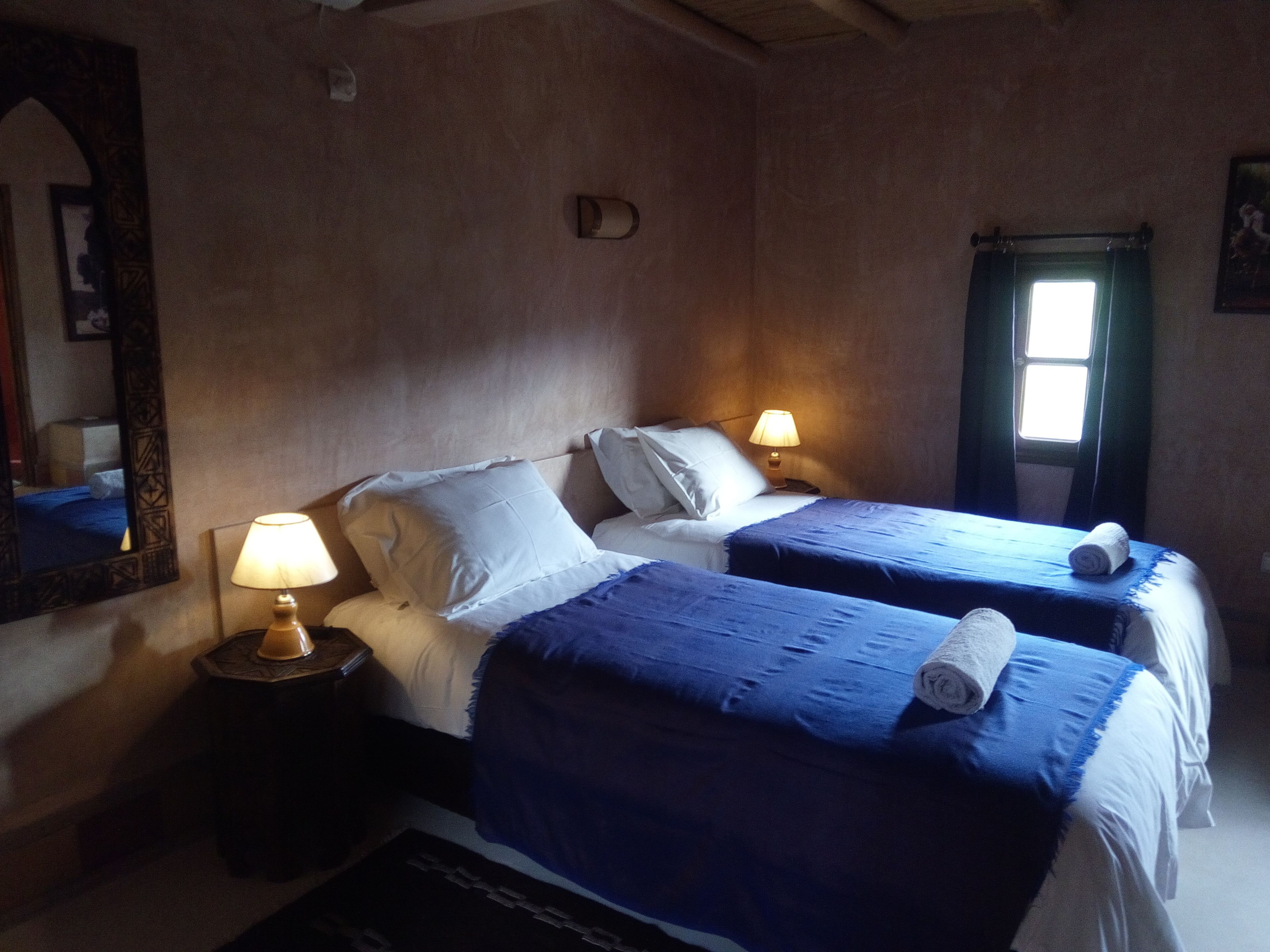 Un dormitorio con dos camas, cada una con cubrecama azul y almohadas blancas. Hay dos lámparas de noche sobre mesitas de madera. Un espejo grande está en la pared izquierda, y una ventana con cortinas en la derecha.