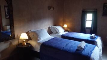Un dormitorio con dos camas, cada una con cubrecama azul y almohadas blancas. Hay dos lámparas de noche sobre mesitas de madera. Un espejo grande está en la pared izquierda, y una ventana con cortinas en la derecha.