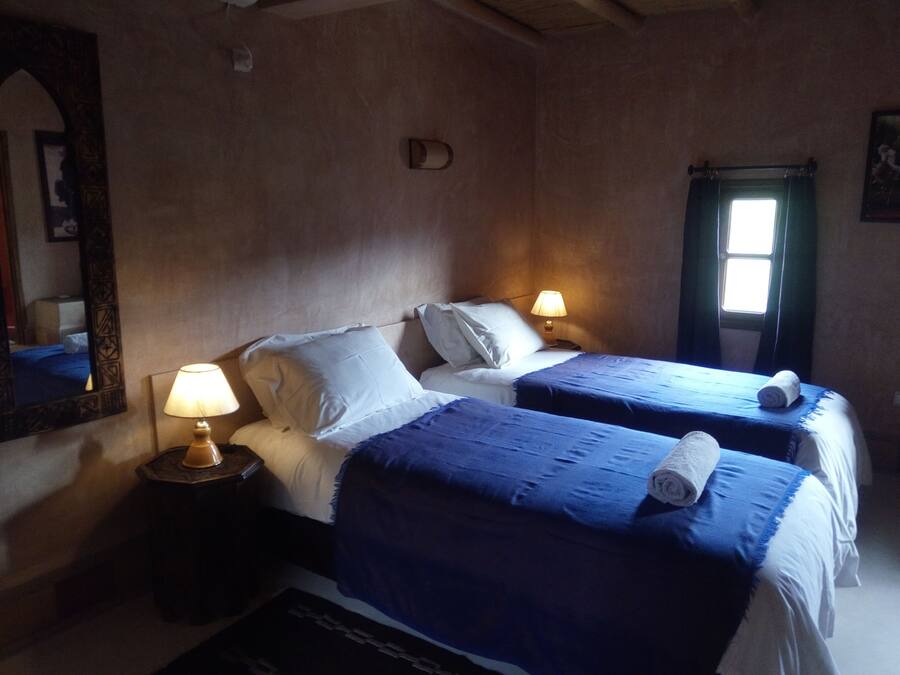 Un dormitorio con dos camas, cada una con cubrecama azul y almohadas blancas. Hay dos lámparas de noche sobre mesitas de madera. Un espejo grande está en la pared izquierda, y una ventana con cortinas en la derecha.