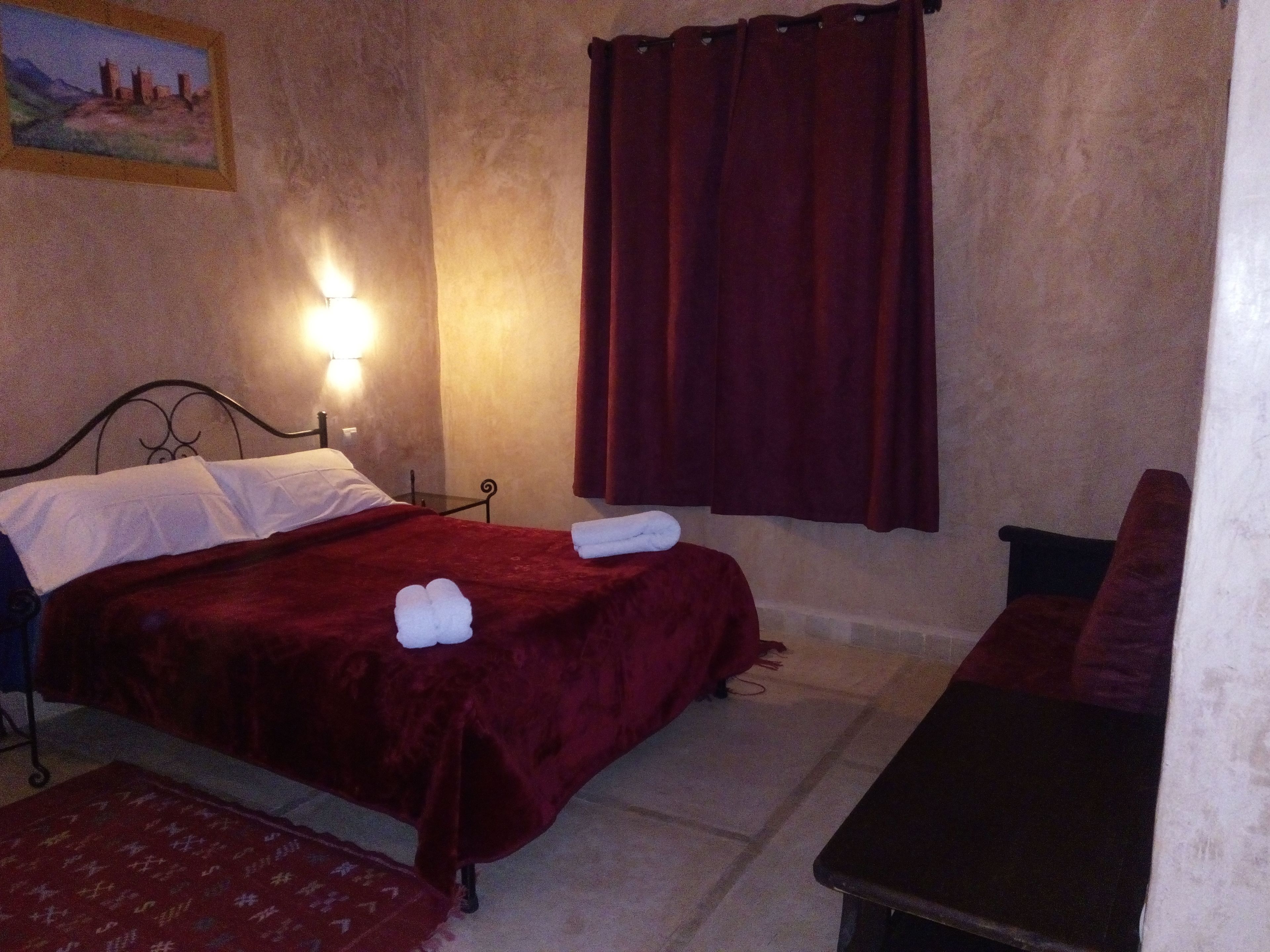 A room with a bed, a small table, a chair, a wall-mounted light, and a window with curtains.