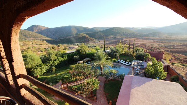 Uitzicht vanaf een balkon op een aangelegde tuin met een zwembad, palmbomen en op de achtergrond bergen.