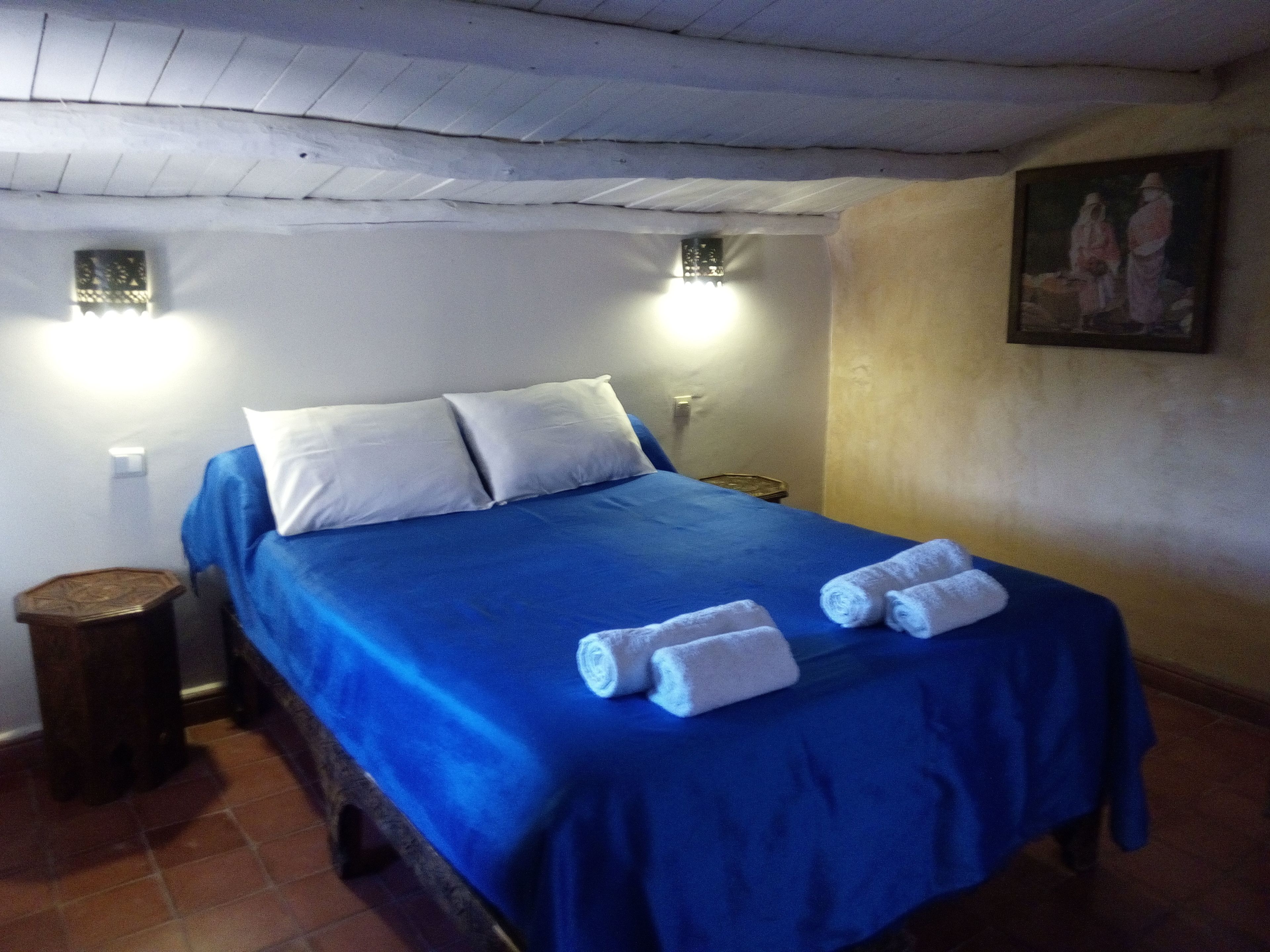 A room with a bed, two wall-mounted lights, a painting, and a small wooden stool.