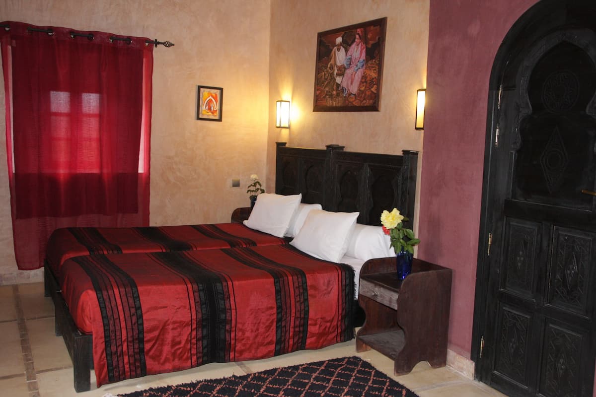 A hotel room with a large bed, a nightstand with a vase of flowers, a wall-mounted painting, and a dark wooden door.