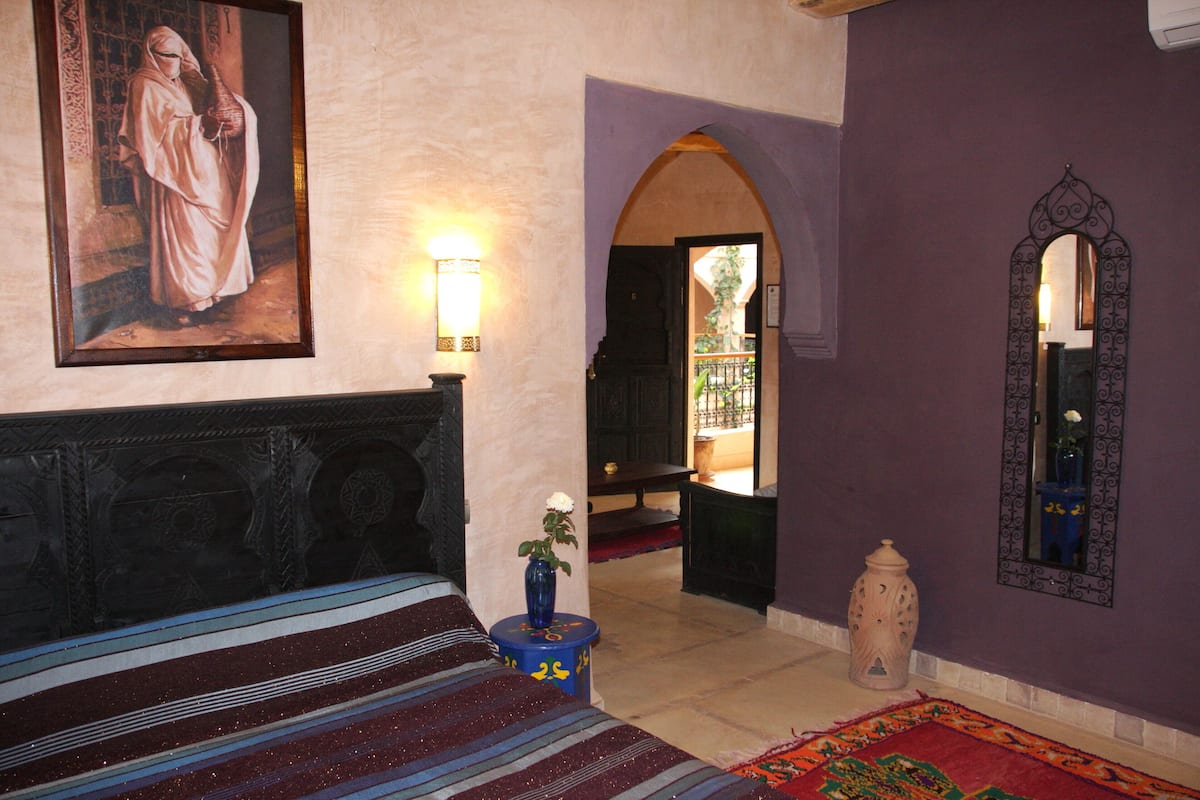 A room with a bed, a wall-mounted painting, a vase with a flower, a mirror, and a door leading to another room.