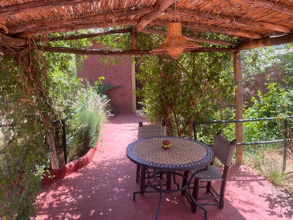 A covered outdoor seating area with a round table and chairs, surrounded by lush greenery.