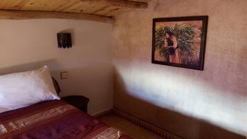 A bedroom with a wooden ceiling, a bed with a red blanket, a small bedside table, and a framed picture on the wall.