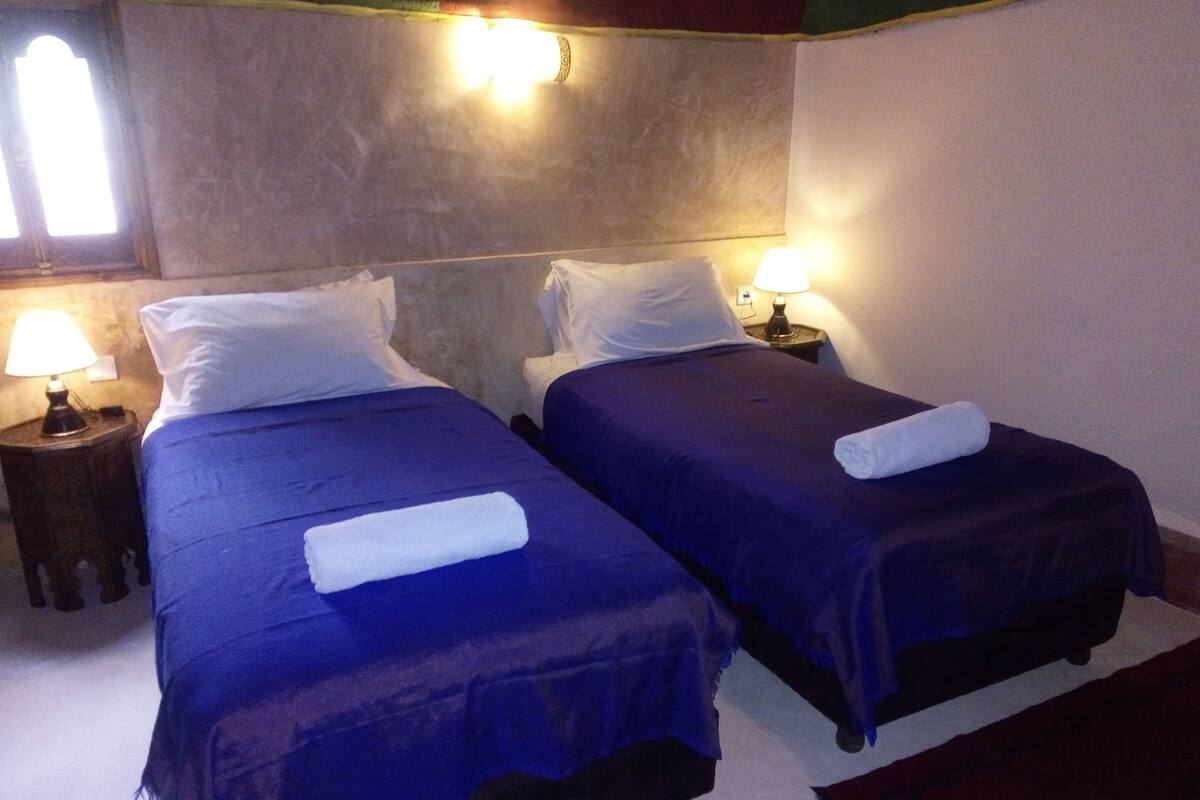 Two beds with blue bedding and white pillows, a nightstand with a lamp, and a window with curtains.