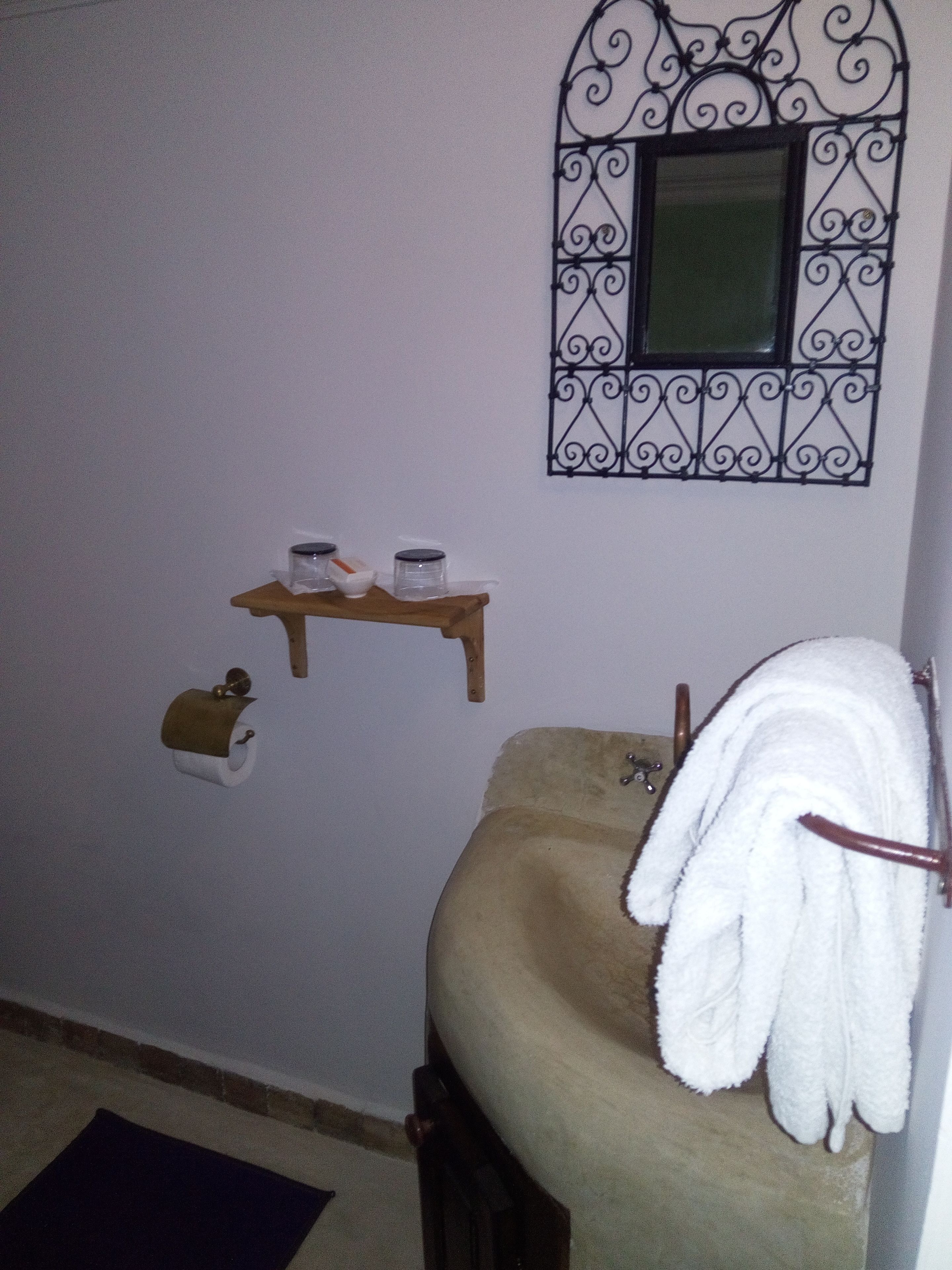 Une salle de bain avec un lavabo beige, un porte-serviettes, un support à papier toilette, une étagère avec deux pots et une grille décorative en métal sur la fenêtre.