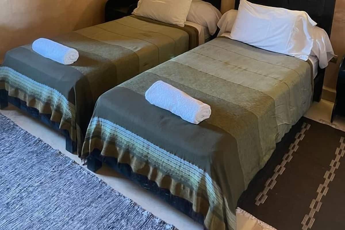 A room with two beds, each with a headboard and a bedside lamp. There are paintings on the walls and a rug on the floor.