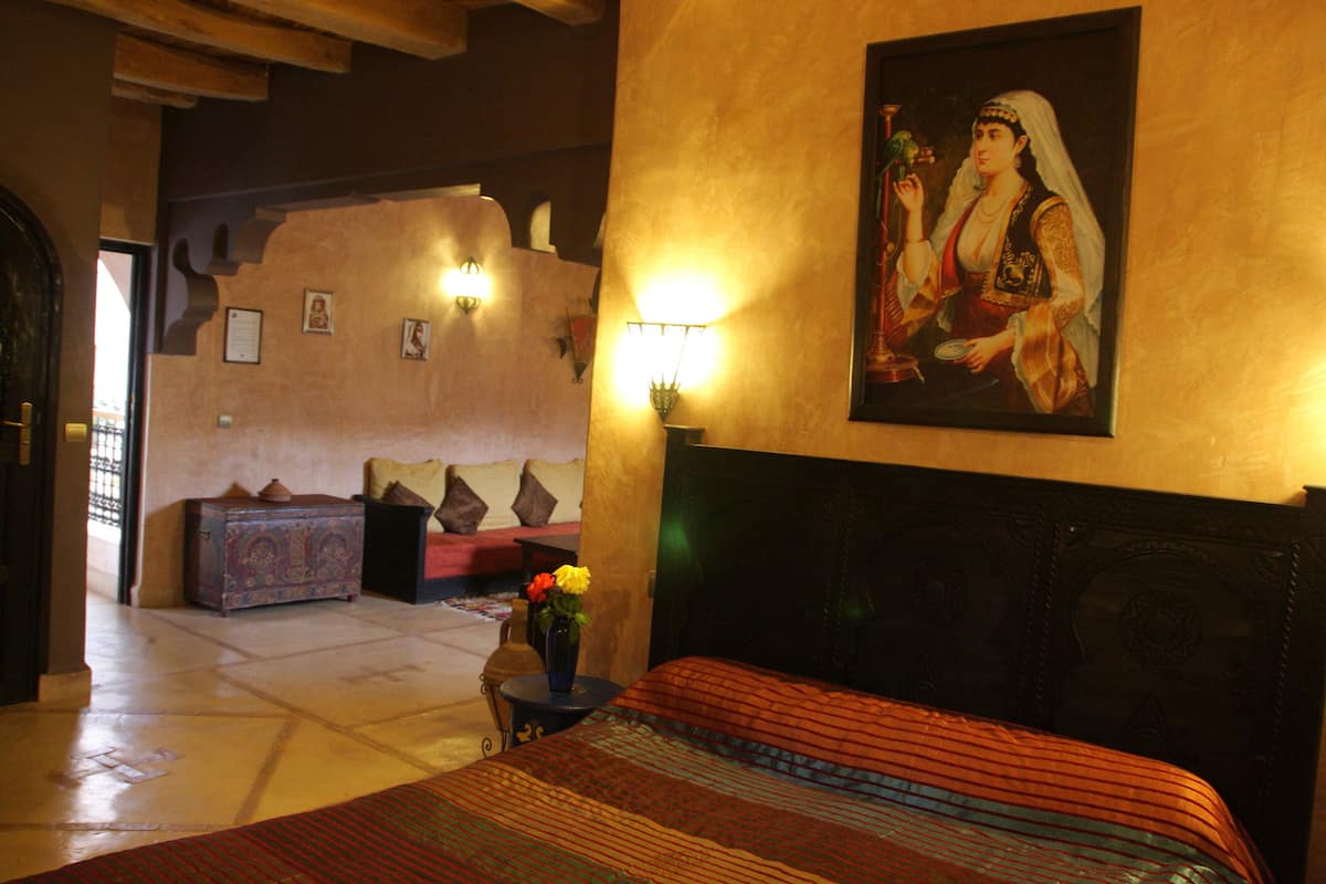A hotel room with a large bed, a nightstand with a vase of flowers, a painting on the wall, and a view of the lobby area.