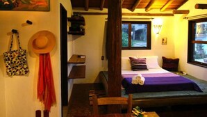 A cozy room with a bed, a chair, a table, a hat, a bag, and a painting on the wall. - Pousada Gopak (Imbituba)