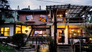 A two-story building with a wooden exterior, a covered outdoor seating area, and a staircase leading to the entrance. - Pousada Gopak (Imbituba)