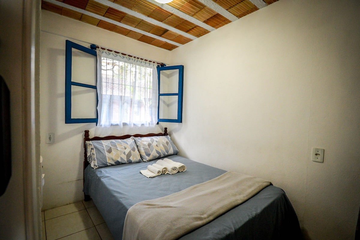 Une petite chambre à coucher avec un lit simple, une fenêtre aux cadres bleus et un rideau.
