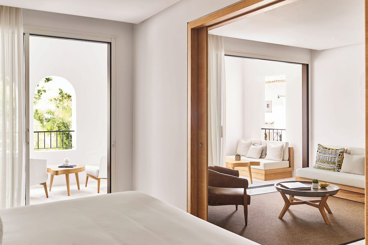 Una habitación de hotel moderna con una cama grande, una mesita redonda pequeña y un sofá. Tiene un balcón con vistas a zonas verdes y una pared blanca.