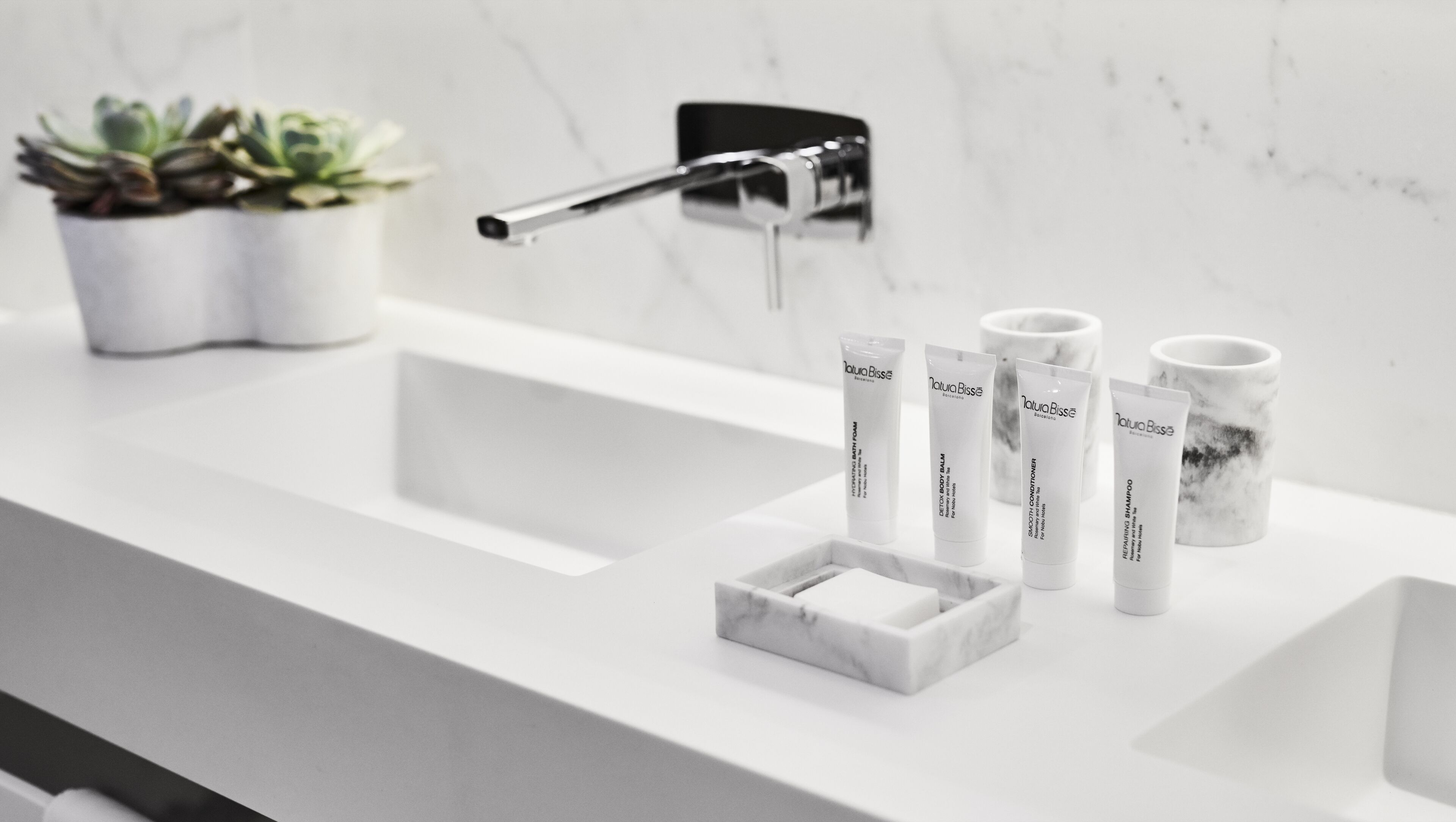 Une salle de bain avec un lavabo blanc, un robinet et trois tubes de produits de la marque Natura Bisse.