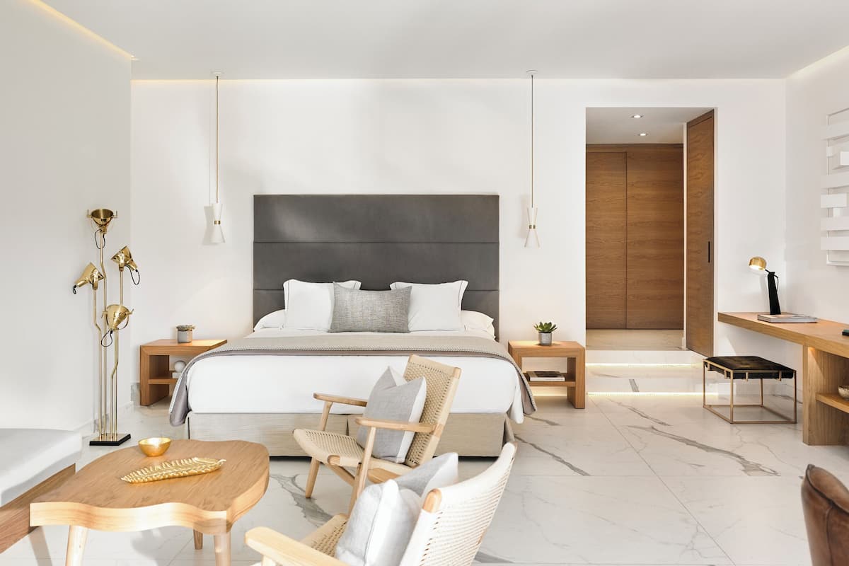 Una habitación de hotel moderna con una cama grande, una zona de estar con una mesa y sillas, un escritorio con silla, y una mesita auxiliar con una planta.
