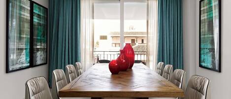 A modern dining room with a large wooden table, white upholstered chairs, and red vases.