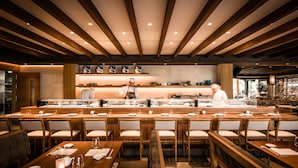 Un ristorante moderno con tavoli e sedie in legno, un bancone centrale con uno chef e vista sulla cucina.