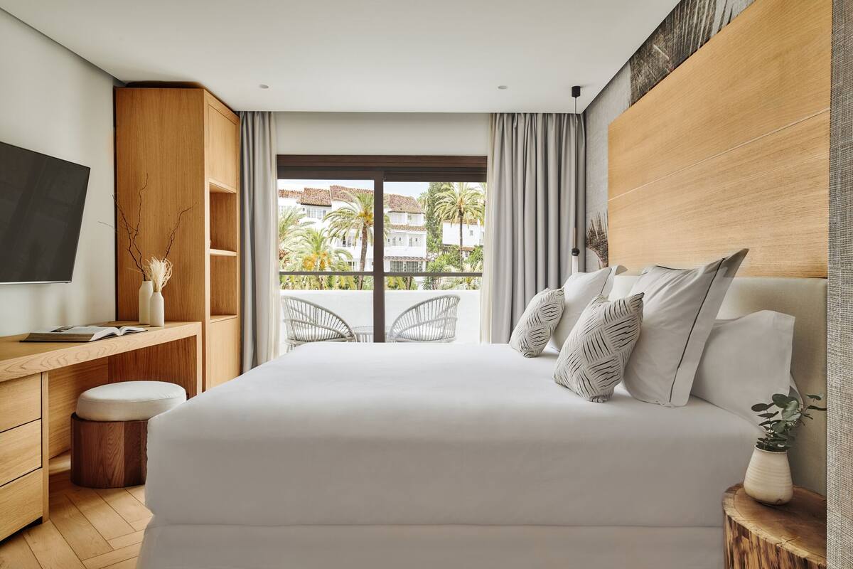 Una habitación de hotel moderna con una cama grande, un escritorio de madera, un televisor y un balcón con vistas a palmeras y edificios.