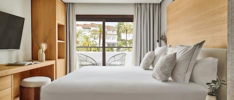 A modern hotel room with a large bed, a wooden desk, a TV, and a balcony with a view of palm trees and buildings.