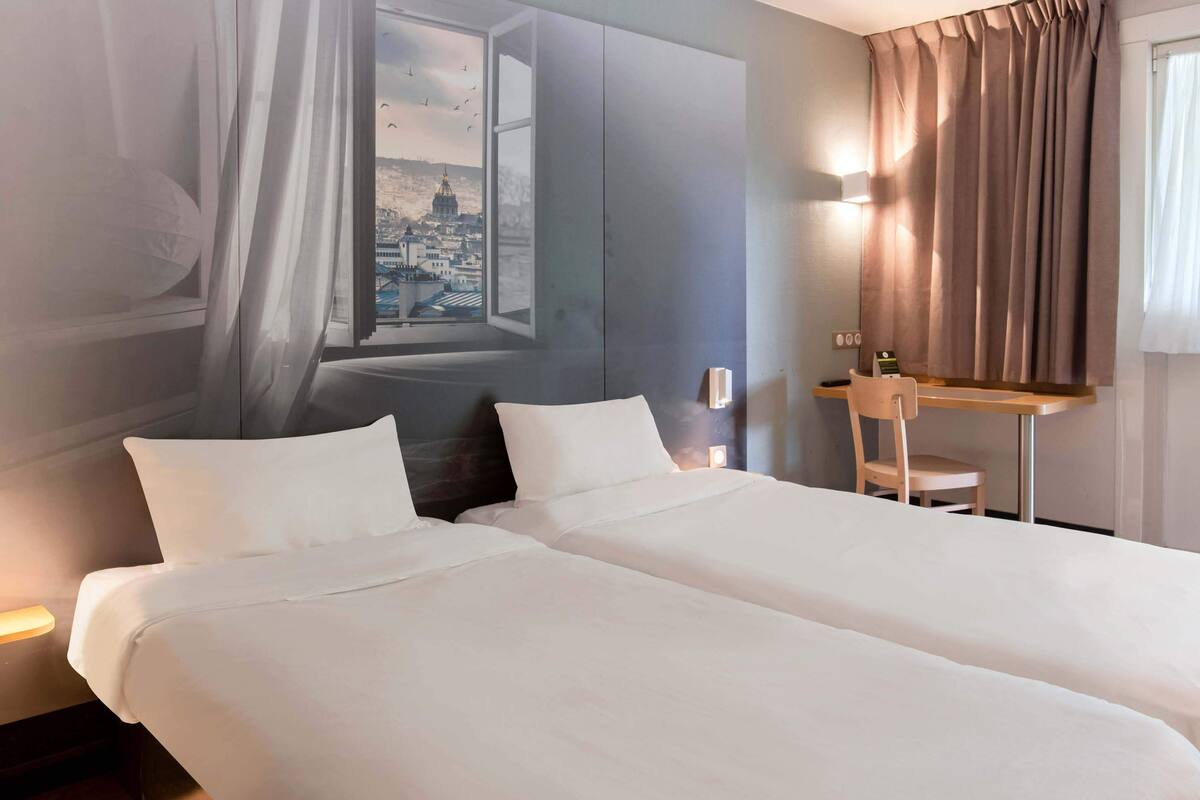 Habitación de hotel con dos camas, un escritorio y vista a un paisaje urbano a través de una ventana.