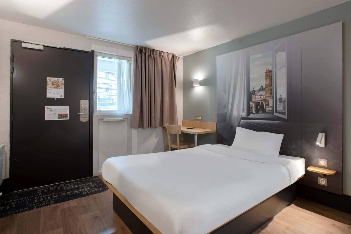Habitación de hotel con cama, escritorio, silla, ventana con cortinas, puerta y una lámpara fijada a la pared.
