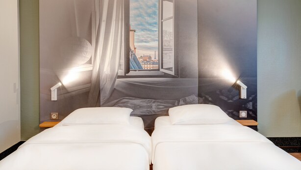 A hotel room with a bed, a nightstand, a wall-mounted lamp, and a view of a cityscape through a window.