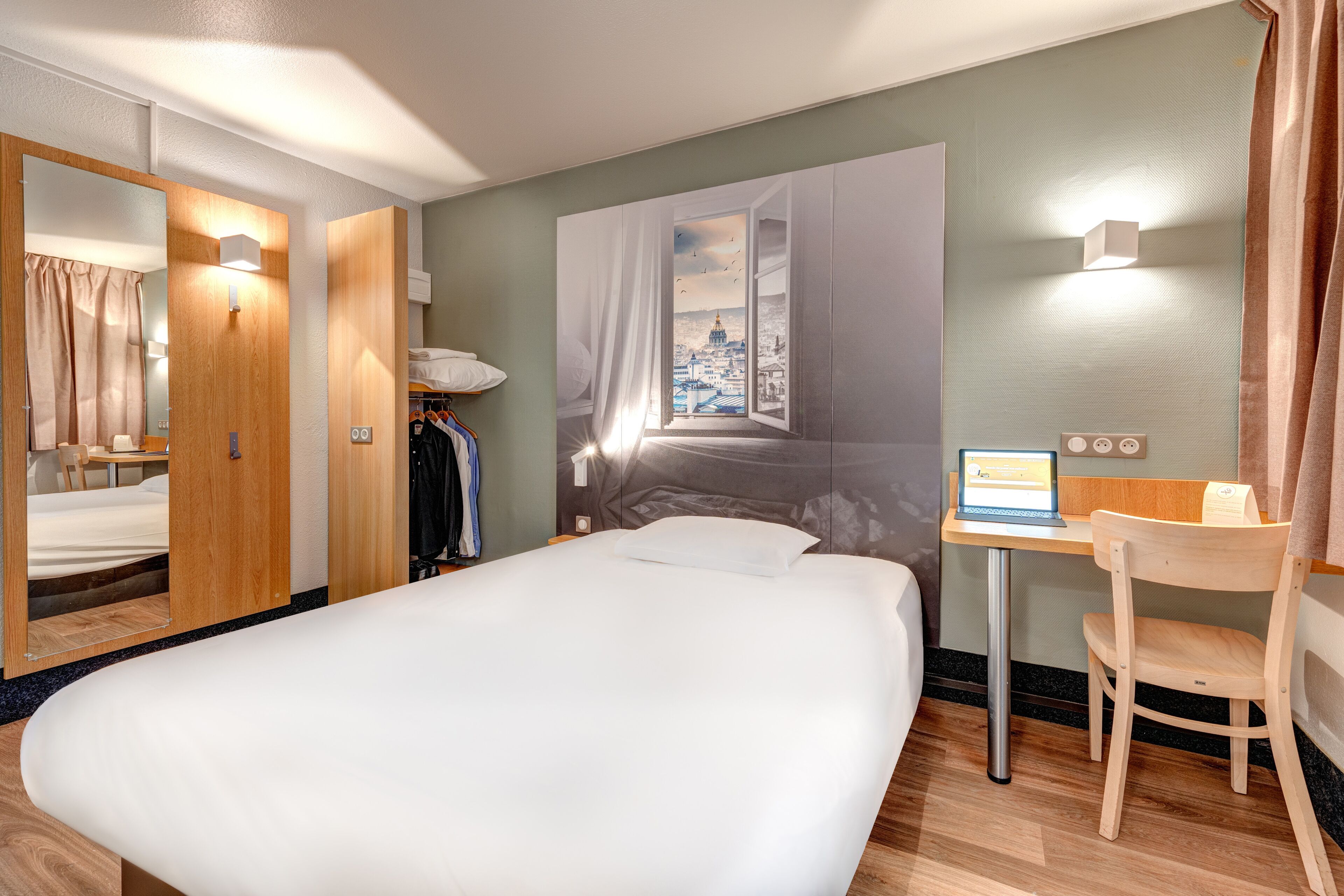 Quarto de hotel com uma cama grande, uma escrivaninha com um laptop, uma cadeira, um guarda-roupa e vista para a paisagem urbana por uma janela aberta.