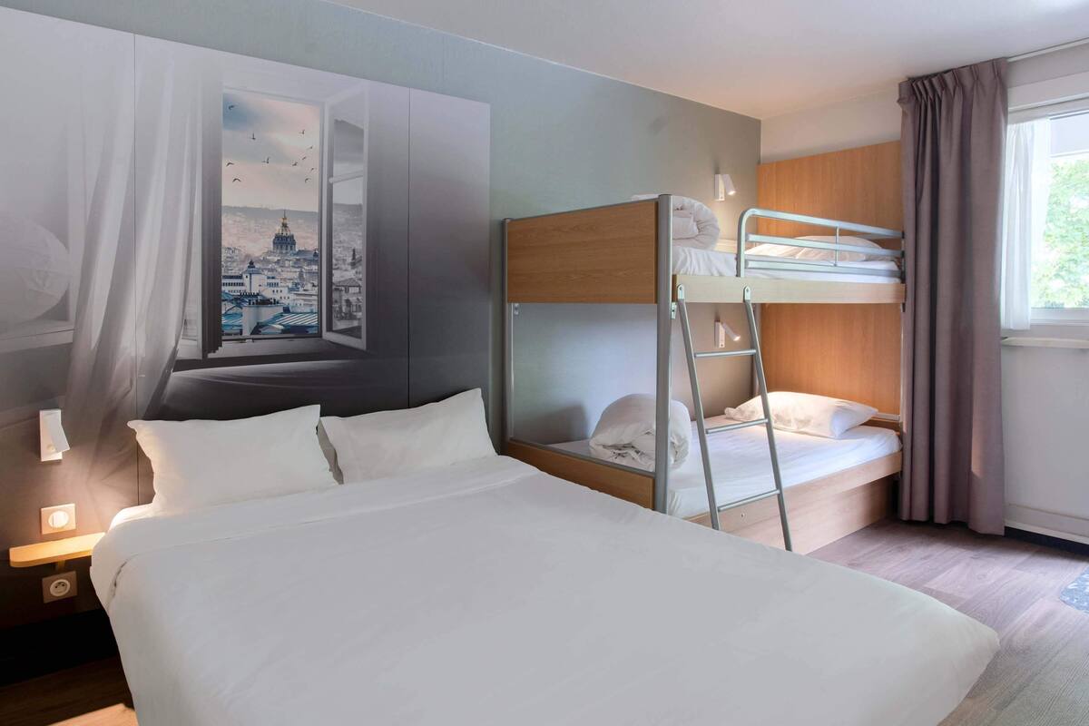 Habitación de hotel con una cama grande, una litera, un mural de paisaje urbano y una ventana con cortinas.