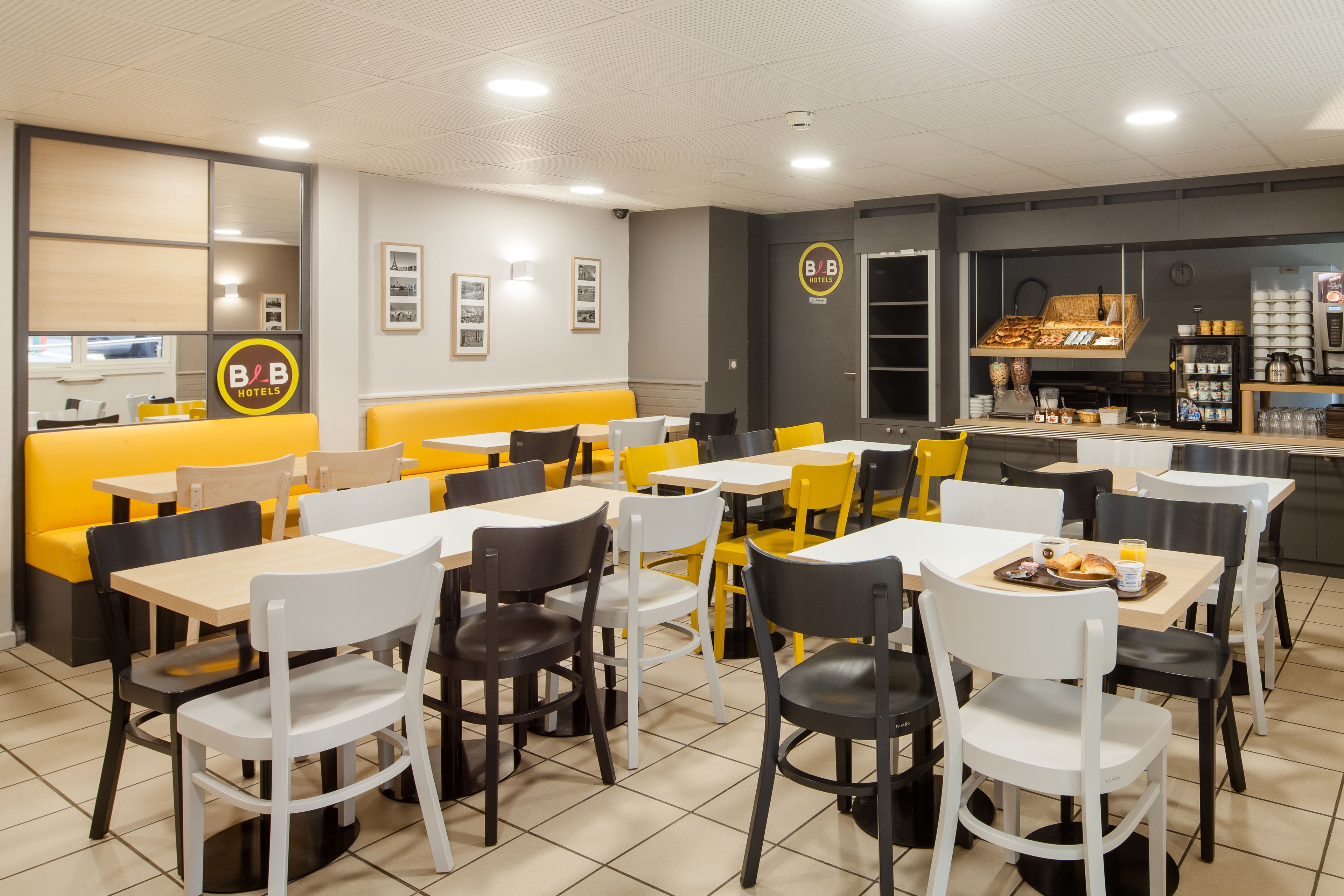 A B&B hotel's dining area with yellow and white seating, a coffee station, and framed pictures on the wall.