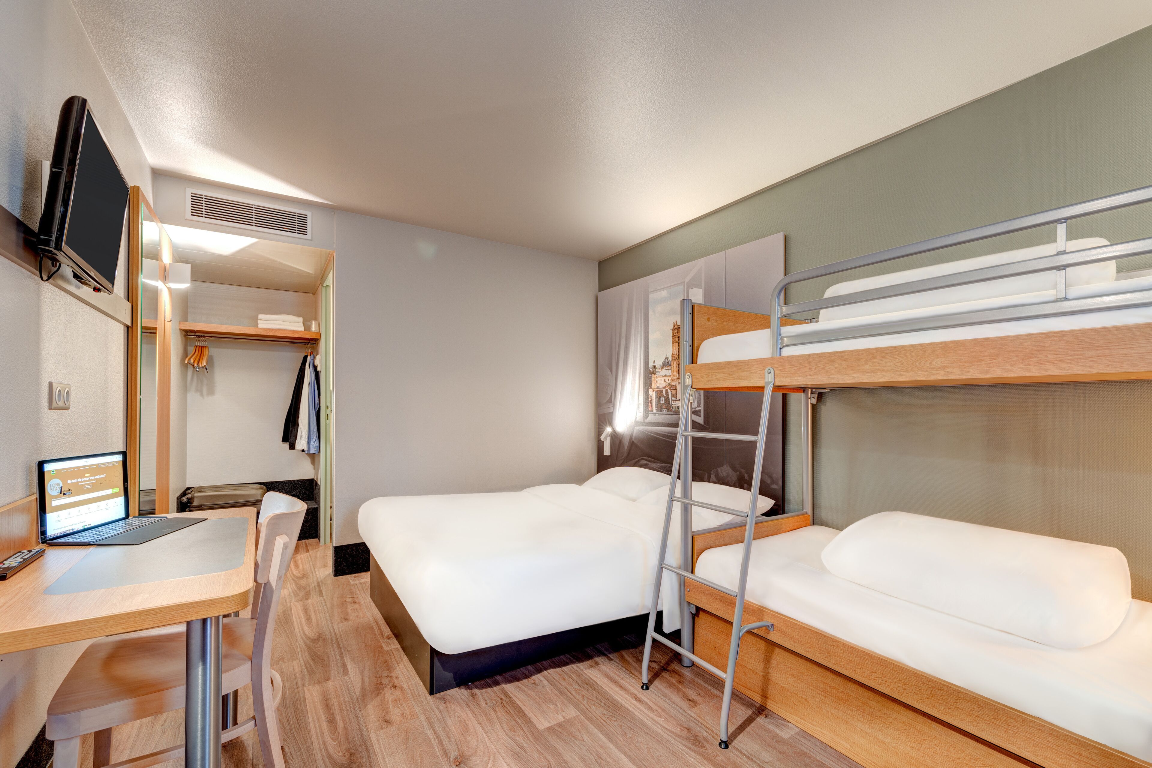 A compact hotel room with two bunk beds, a desk with a laptop, a TV mounted on the wall, and a small closet.