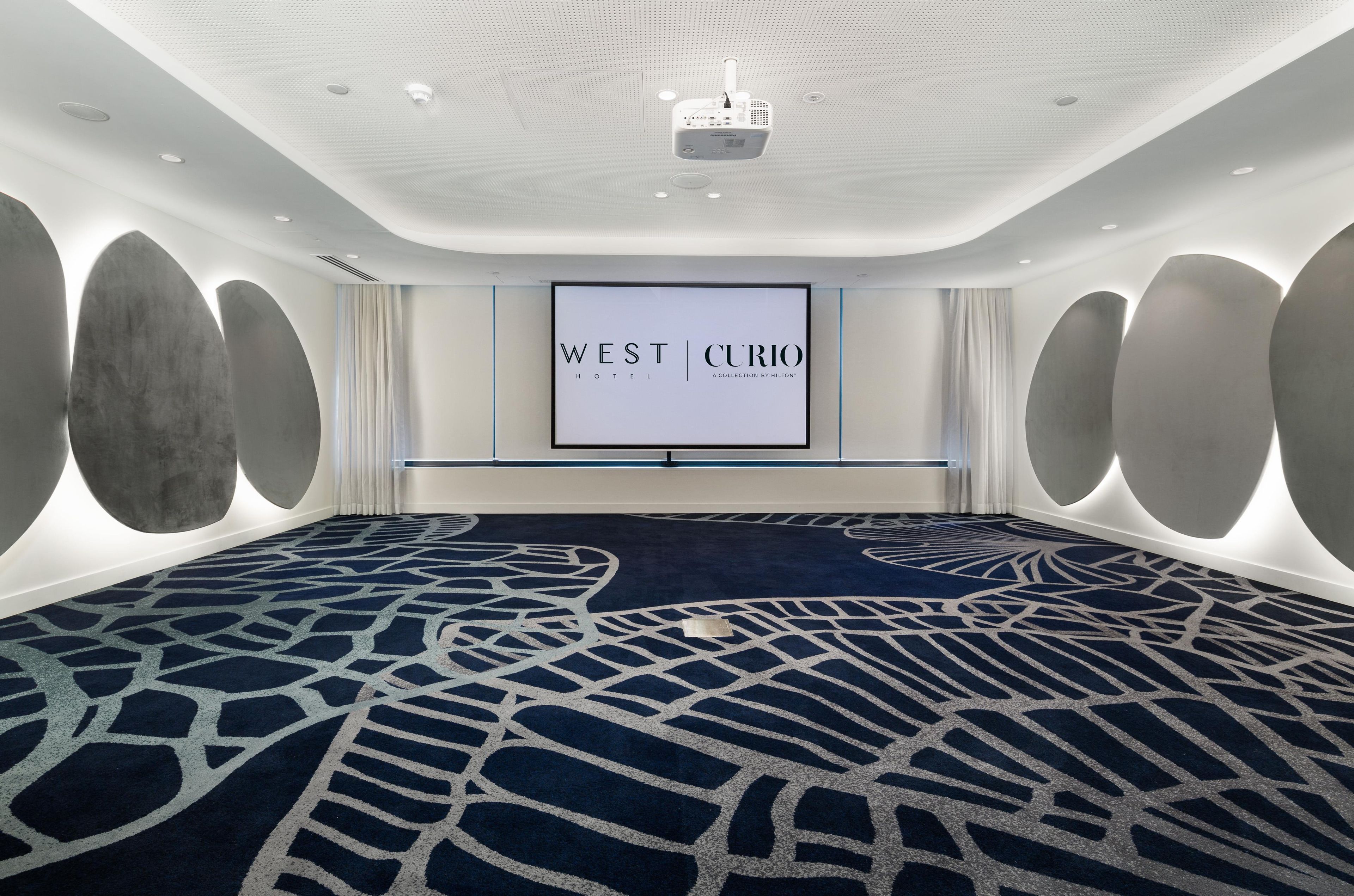 Une grande salle de conférence dotée d’un tapis à motifs sombres, d’un grand écran affichant le logo de l’hôtel « WEST CURIO », et d’un projecteur fixé au plafond.