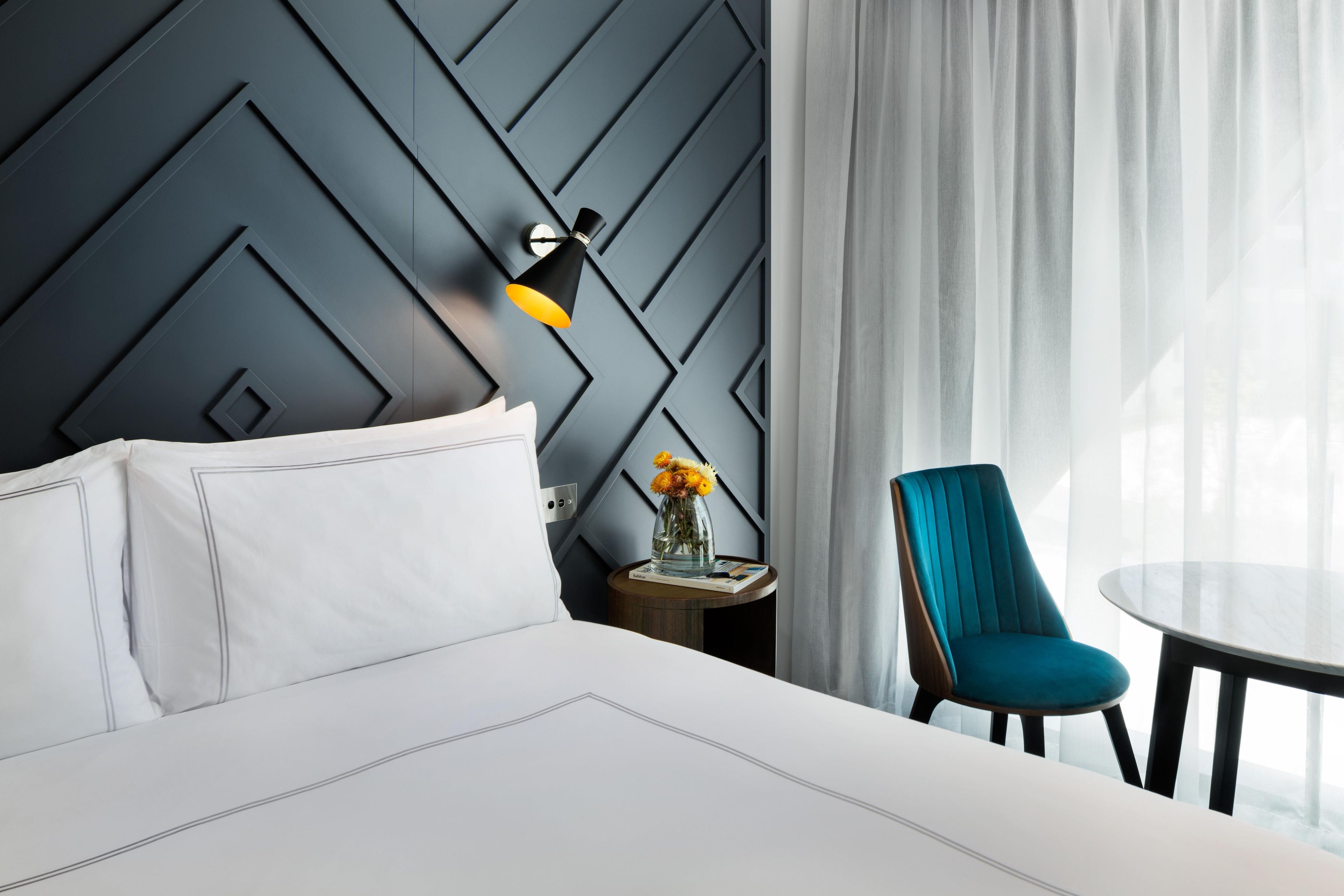 Quarto de hotel moderno com cama, uma cadeira, uma mesa e uma luminária fixada na parede.