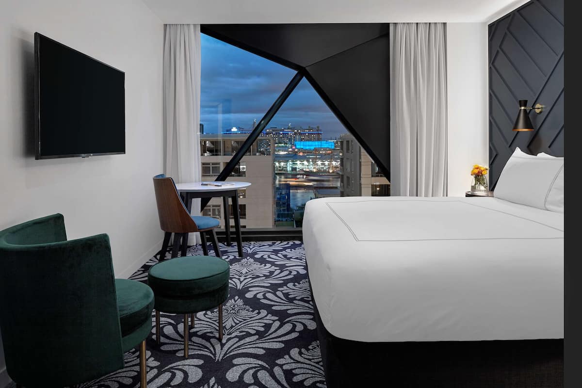 A hotel room with a large bed, a desk, a chair, and a TV. There is a view of a cityscape through a window.