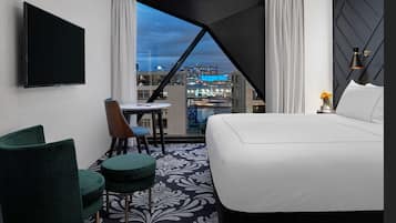 A hotel room with a large bed, a desk, a chair, and a TV. There is a view of a cityscape through a window.