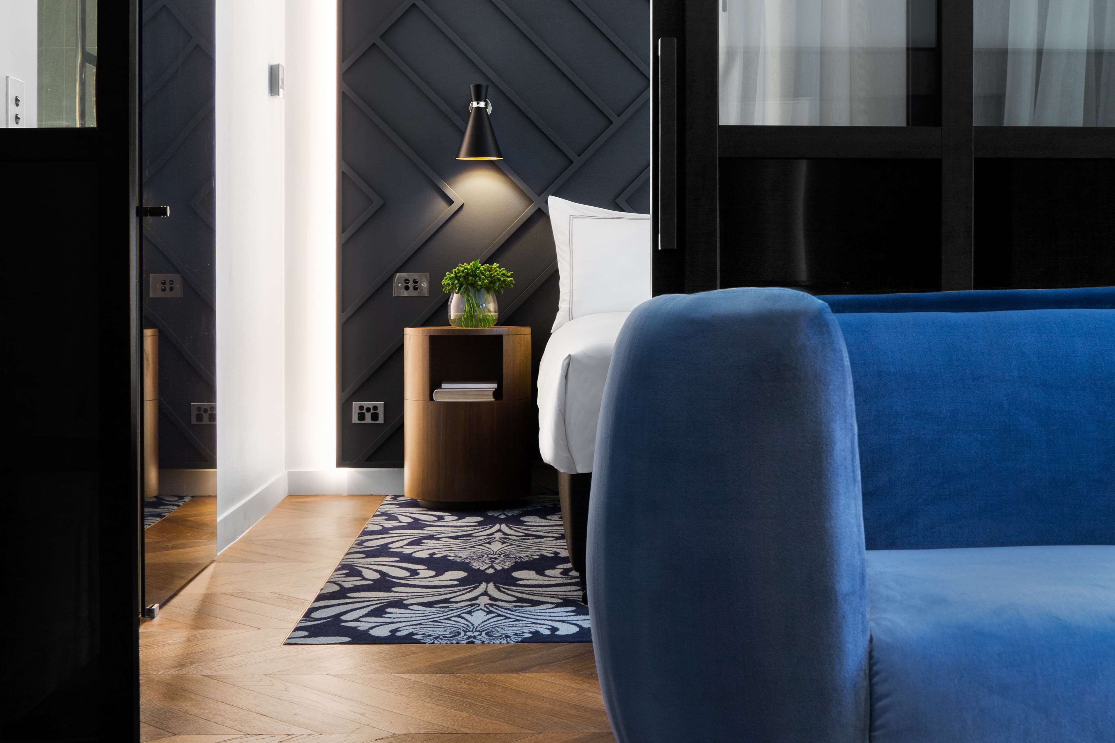 Una habitación de hotel moderna con un sofá azul, un tapete estampado, una mesita de madera con una planta y una pared con diseños geométricos.