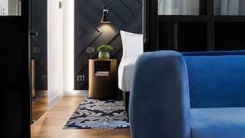 Una habitación de hotel moderna con un sofá azul, un tapete estampado, una mesita de madera con una planta y una pared con diseños geométricos.