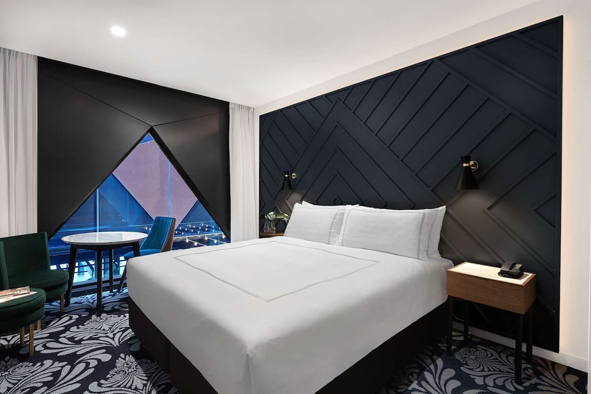 A modern hotel room with a large bed, a small table, a chair, and a view of the cityscape through a window.