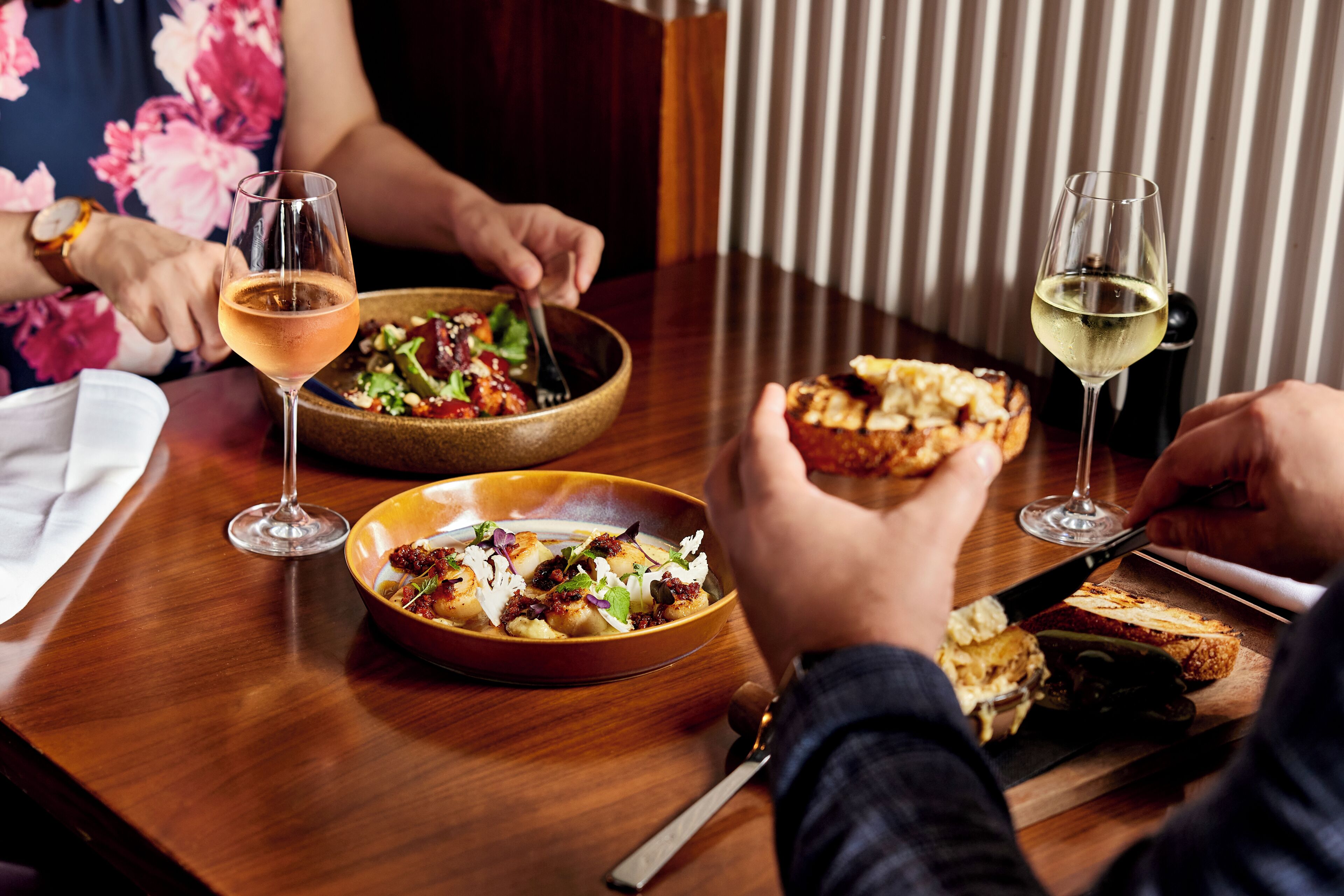 A dining table with two bowls of food, a glass of wine, and a person cutting bread.