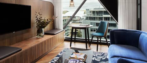 A modern living room with a flat-screen TV, a blue sofa, a coffee table with books, a vase with plants, and a city view through large windows.