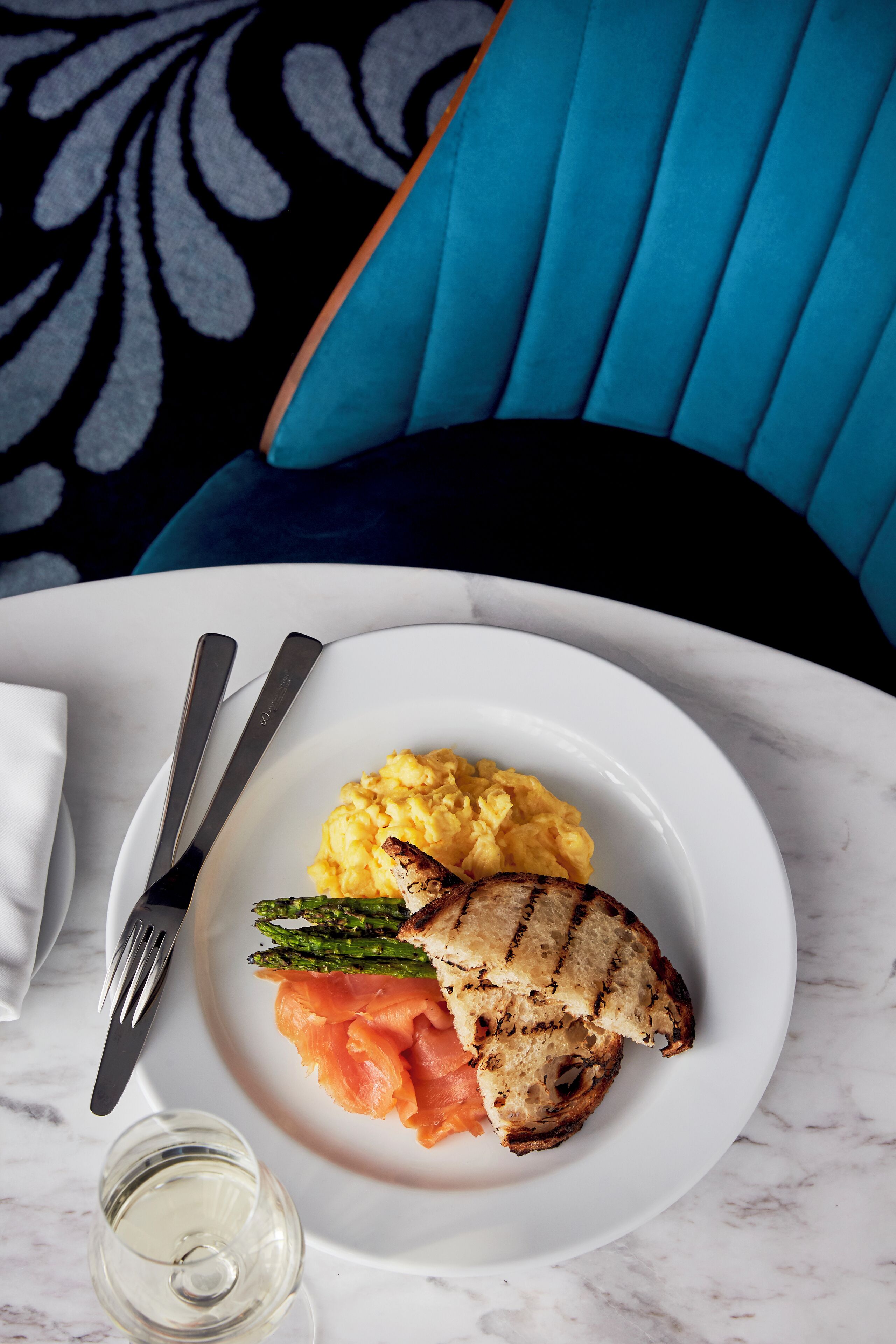 A plate with grilled chicken, asparagus, and salmon on a white plate, accompanied by a fork and knife, and a glass of white wine.
