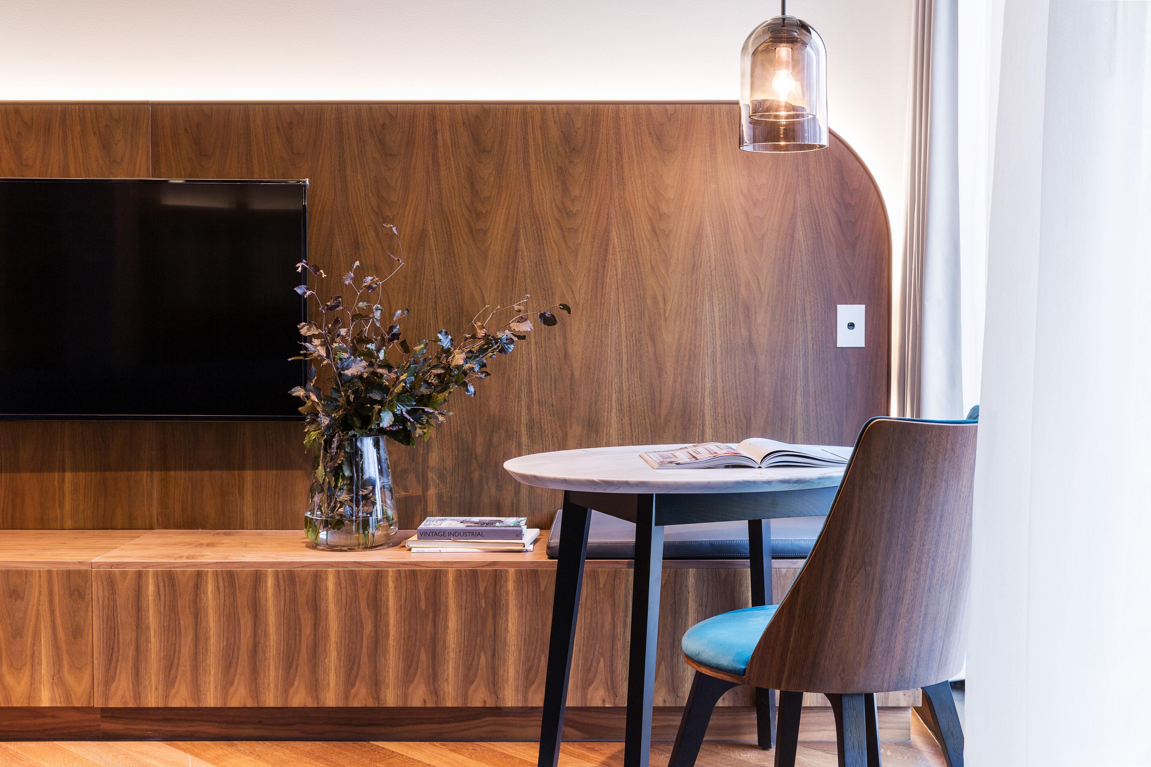 Un espacio moderno para vivir con un televisor de pantalla plana montado en una pared de madera, una mesa redonda con una sola silla y un jarrón con flores secas sobre un banco de madera.