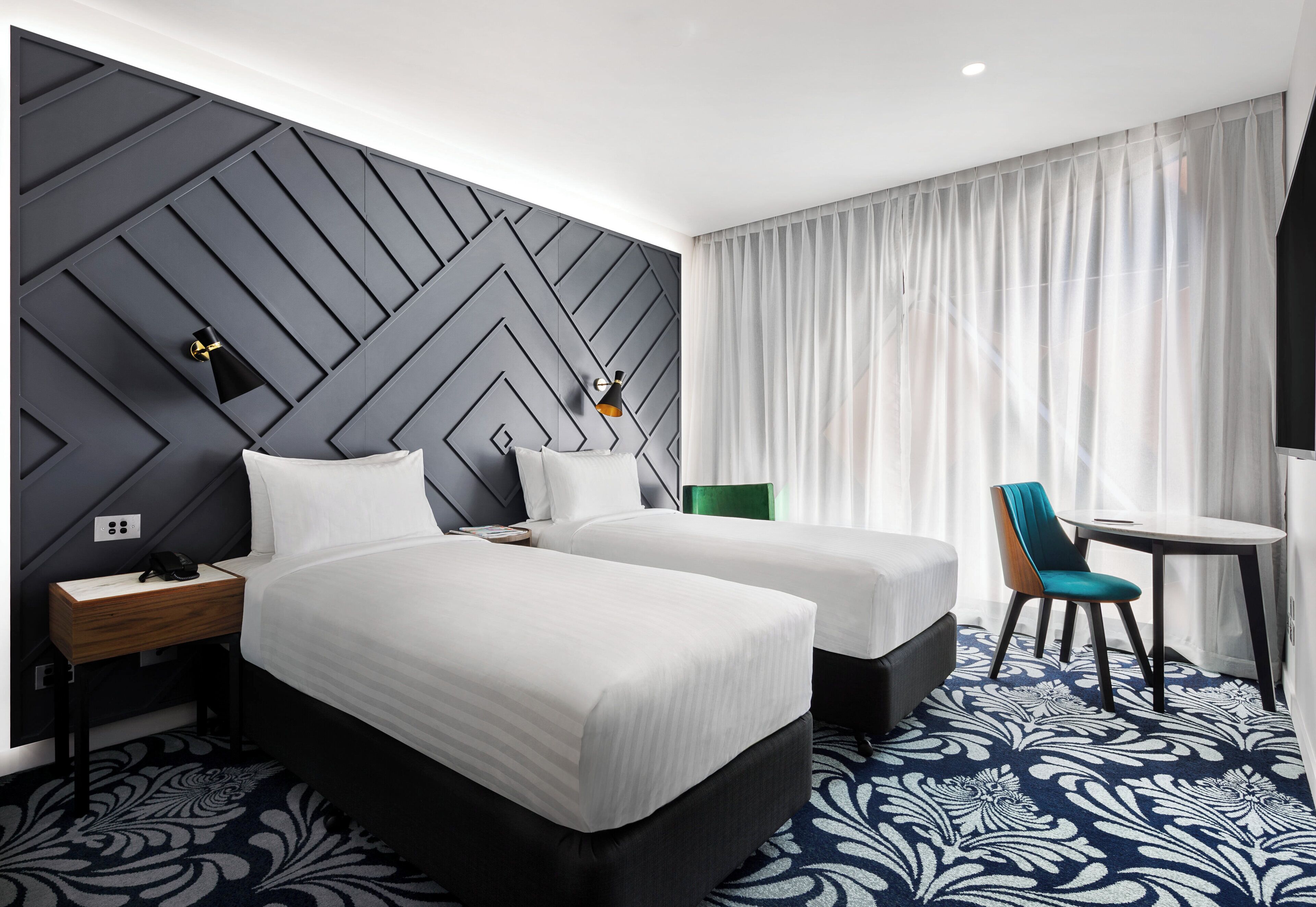 A hotel room with two beds, a desk, a chair, and a large window with curtains.