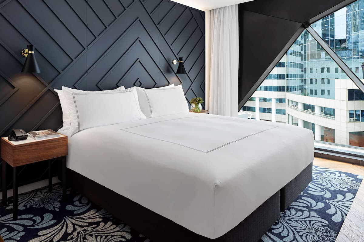 A modern hotel room with a large bed, a nightstand with a lamp, and a view of a cityscape through a large window.